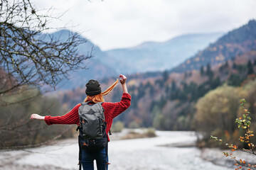 a traveler in a hat, sweater and trousers gestures with her hands on the river bank in the mountains
