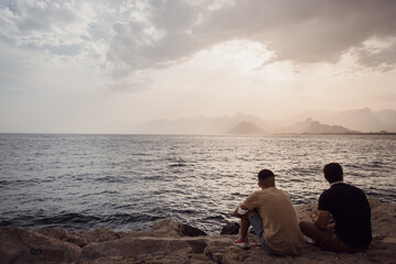 Fototapeta premium Summer landscape overlooking the Mediterranean coast. Layers of mountains on the horizon, rays of the sun breaking through the clouds. Two men sit and watch the sunset. Antalya, Turkey