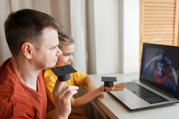Father with kid talking with eldest son wearing graduation gown and cap and cheers him with university graduation, they made small paper cap for this online video call, distant education