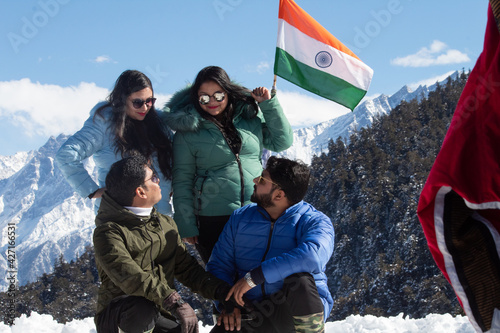 Four friends looking at each other while posing with Indian flag in front of snowcapped mountain