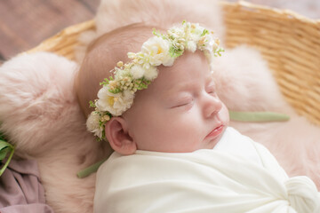 Baby girl in a wicker basket of vine decorated with  pink fur in a light winding and a flower wreath on her head. Spring photo. Flowers and children. Happy motherhood 