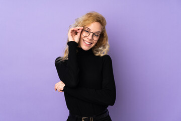 Young blonde woman isolated on purple background with glasses and happy