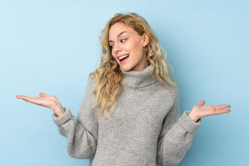 Young blonde woman wearing a sweater isolated on blue background holding copyspace with two hands