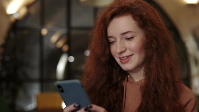 Crop view of pretty woman laughing while using smartphone and sitting in cafe. Attractive female person with red curly hair chatting in social media and having good mood.