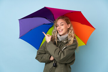 Young blonde woman holding an umbrella isolated on blue background and pointing it
