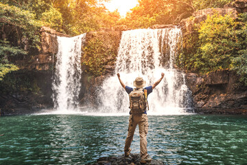Happy man backpacker enjoying amazing tropical waterfall raised hands .