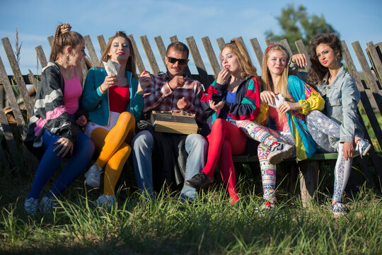 A group of people dressed in the style of the nineties sit on a bench by a skewed fence.