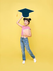 Happy Asian cute schoolgirl with graduation hat and book isolated on yellow background.
