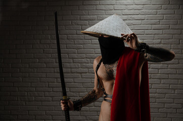 samurai man in a red cape and an Asian triangular hat with tattoos and a katana sword in his hands against the background of a brick wall