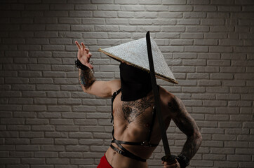 samurai man in a red cape and an Asian triangular hat with tattoos and a katana sword in his hands against the background of a brick wall