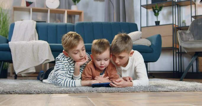 Handsome peaceful friendly three brothers relaxing on fluffy carpet and watching exciting cartoon or show on smartphone