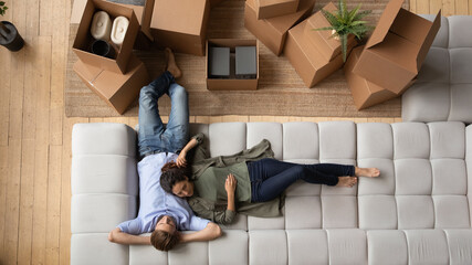 Top view happy couple tenants relaxing lying on couch after relocation in first own apartment, young man and woman resting on moving day in living room with boxes, mortgage or rent concept