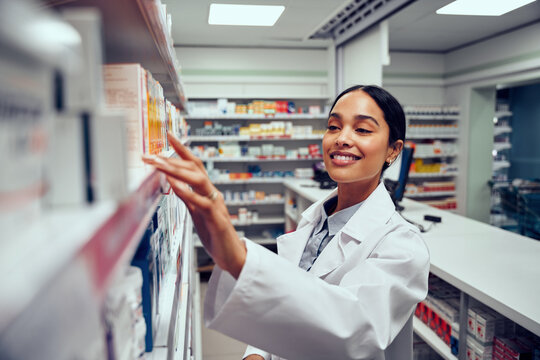 Smiling young female pharmacist wearing labcoat standing behind counter looking for medicine in shelf
