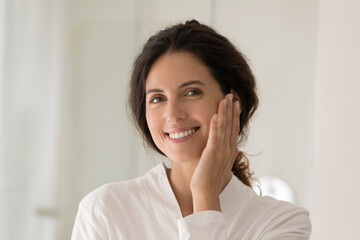 Head shot portrait smiling beautiful woman touching cheek, perfect smooth face skin, looking at camera, happy attractive young female in bathrobe enjoying skincare spa procedure, natural beauty