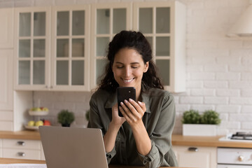 Close up smiling woman using smartphone, typing, browsing apps, sitting at table with laptop in modern kitchen, positive young female browsing mobile device apps, writing message, scrolling