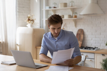 Focused man working with documents, paying bills, sitting at table with laptop in modern kitchen, businessman freelancer reading correspondence, checking, analyzing financial report, paperwork