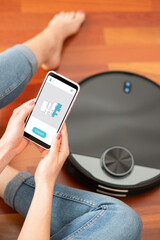 Young girl feet at home using a smart phone to control a round smart automatic vacuum, machine housework robot cleaning the floor