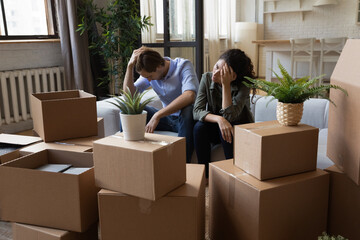 Unhappy frustrated couple sitting on couch with cardboard boxes, eviction, family having problem...