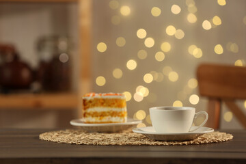 Hot drink in cup and piece of cake on wooden table against blurred lights indoors