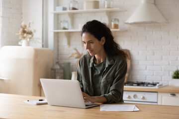 Confident focused woman using laptop, looking at screen, freelancer working on project online at home, young female sitting at table in kitchen, chatting in social networks, studying, browsing apps