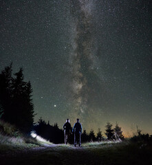 Fototapeta premium Picturesque view of night starry sky over mountain valley with couple travelers. Young woman and man holding hands while standing on mountain road under beautiful sky with stars and Milky Way.