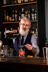 Brutal male bartender with beard prepares alcohol drink with a steel shaker at bar counter