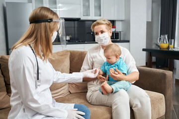 Obraz premium Competent doctor in protective mask and gloves visiting her little patient at home. Young mother sitting on couch with cute toddler i on hands. Medical treatment.