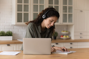 Close up smiling woman in headphones writing taking notes, studying online, young female sitting at table in kitchen with laptop, watching webinar, listening to lecture, involved in training