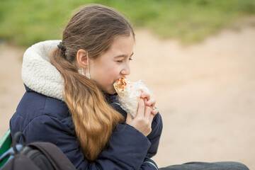 A teenage schoolgirl girl is sitting on a bench with a backpack in a medical mask and eating shawarma