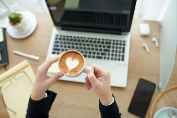 Hands of businesswoman holding cup of latte with heart shape foam after finishing working on laptop