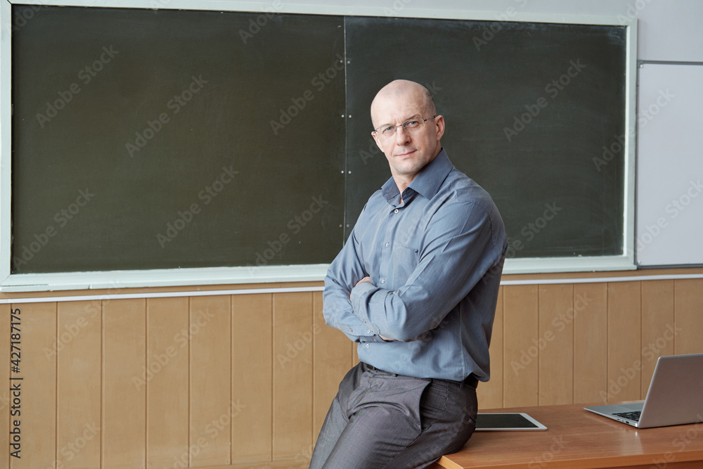 Poster bald professor of university in smart casualwear sitting on desk against blackboard while crossing a