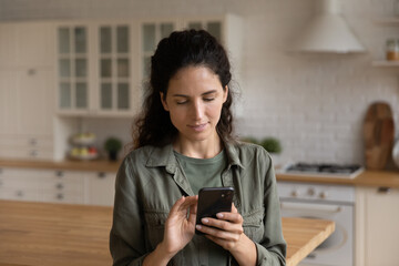 Close up focused woman looking at phone screen, browsing mobile device apps, standing in kitchen at home, confident young female typing, writing message, shopping or chatting online, scrolling