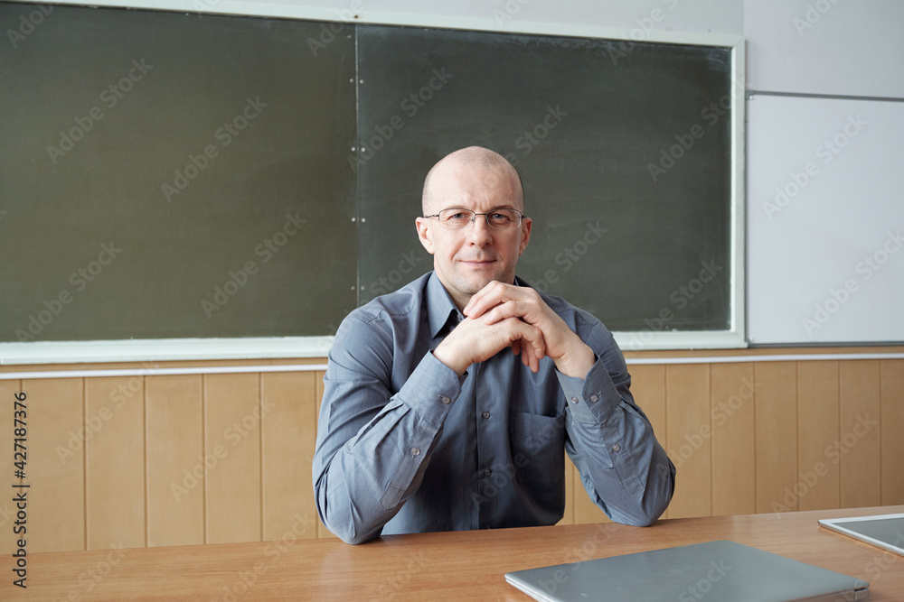 Aufkleber bald professor of modern university sitting by desk against blackboard while keeping hands by chin a