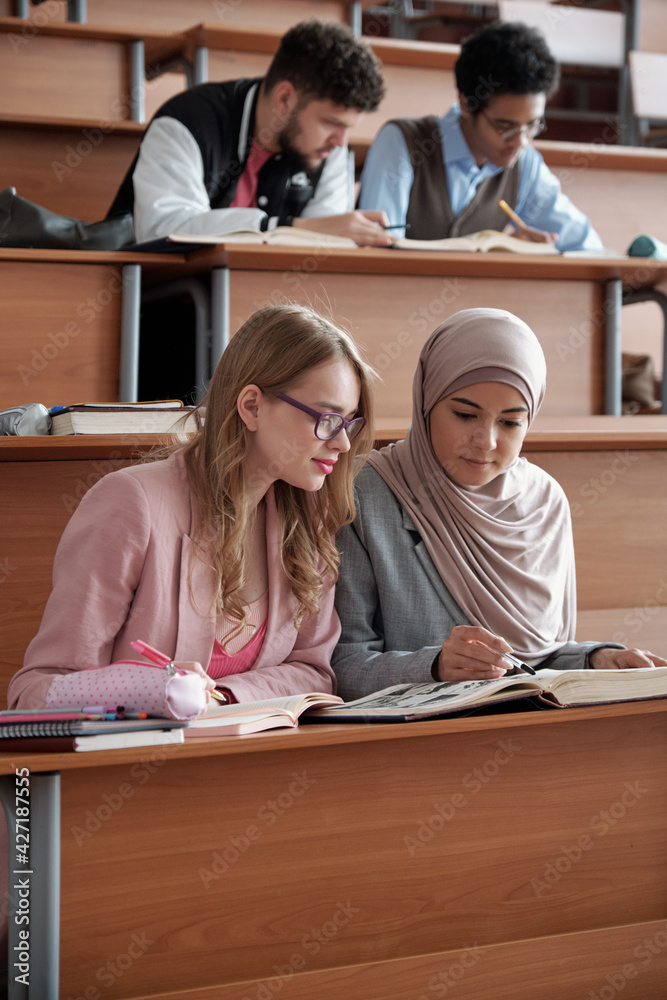 Poster two intercultural female students of college or university sitting on the first row by desk in lectu