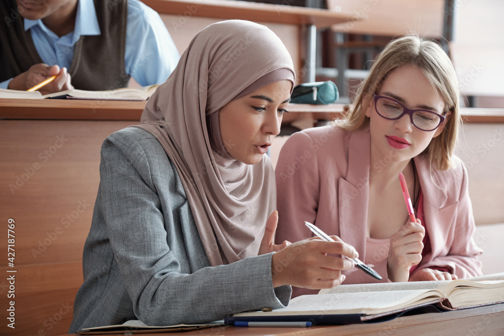Poster Two intercultural female students sitting by desk in lecture hall and discussing passage in book while one of them pointing at page