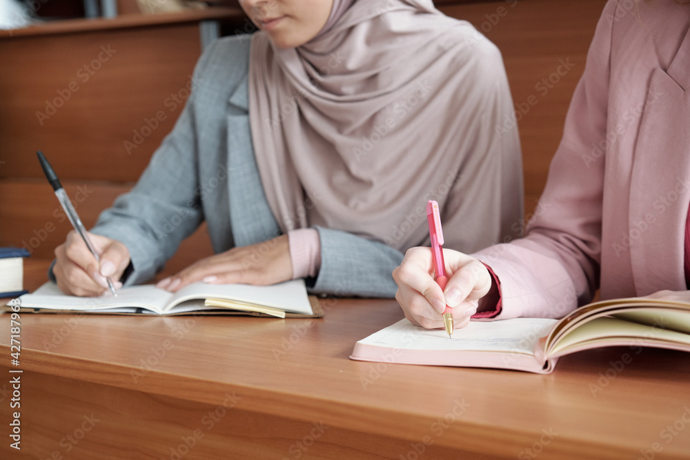 Aufkleber hands of two young female students with pens making notes in copybooks at lecture while sitting in r