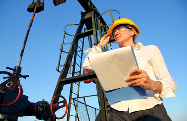 Successful female engineer with a clipboard control the operation of the oil pump, close up of young woman with safety goggles looks at sunset