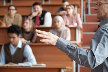 Hand of mature professor in casualwear standing in front of audience and explaining subject of lecture to young intercultural students