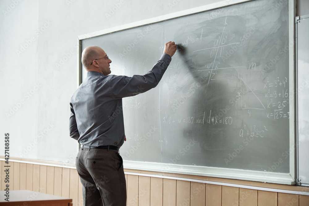 Poster Bald professor of university in smart casualwear standing in front of blackboard and writing down algebraic formula during explanation