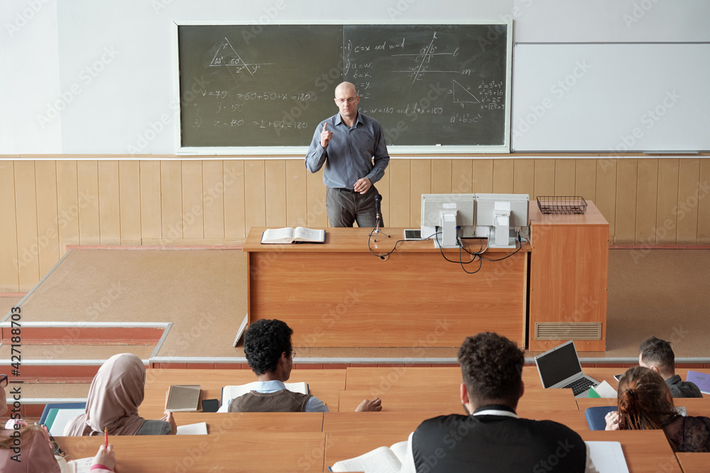 Poster bald mature professor in smart casualwear standing by desk in front of audience and explaining subje