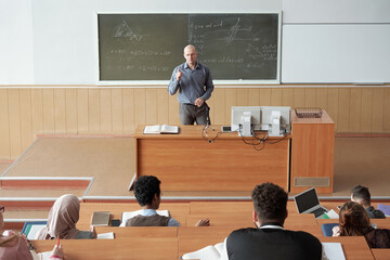 Bald mature professor in smart casualwear standing by desk in front of audience and explaining...