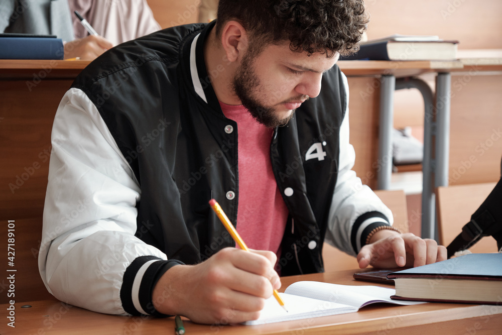 Poster Serious guy in casualwear bending over desk and making notes in copybook while scrolling in smartphone during lecture in university
