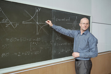 Confident mature professor standing in front of blackboard in lecture hall, pointing at graph and...