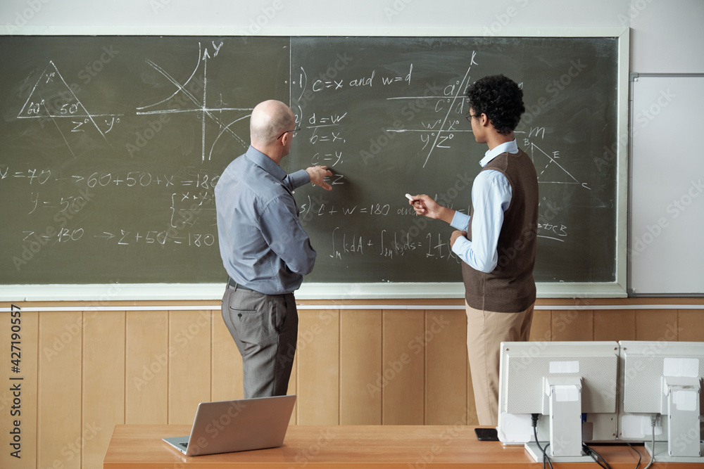 Poster mature bald professor and young mixed-race student discussing solution of equation by blackboard whi