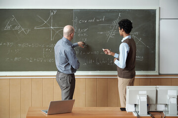 Mature bald professor and young mixed-race student discussing solution of equation by blackboard...