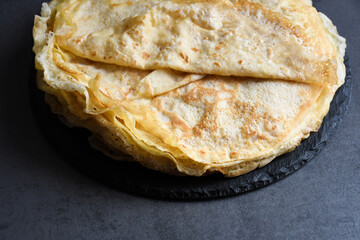 Pancakes, flat and round flour products made from batter by frying in a pan, lie on a black slate plate against a gray background. Pancakes are used as an independent dish and together with fillings.