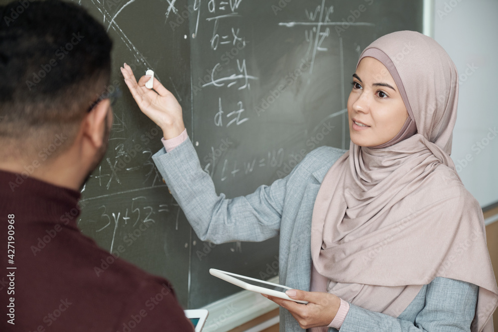 Poster Young female student in hijab looking at professor while both standing by blackboard and girl pointing at graph while solving equation at lesson