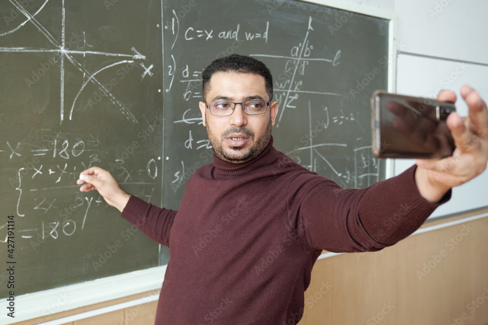 Leinwandbilder mixed-race male in casualwear holding smartphone in front of himself while standing by blackboard an