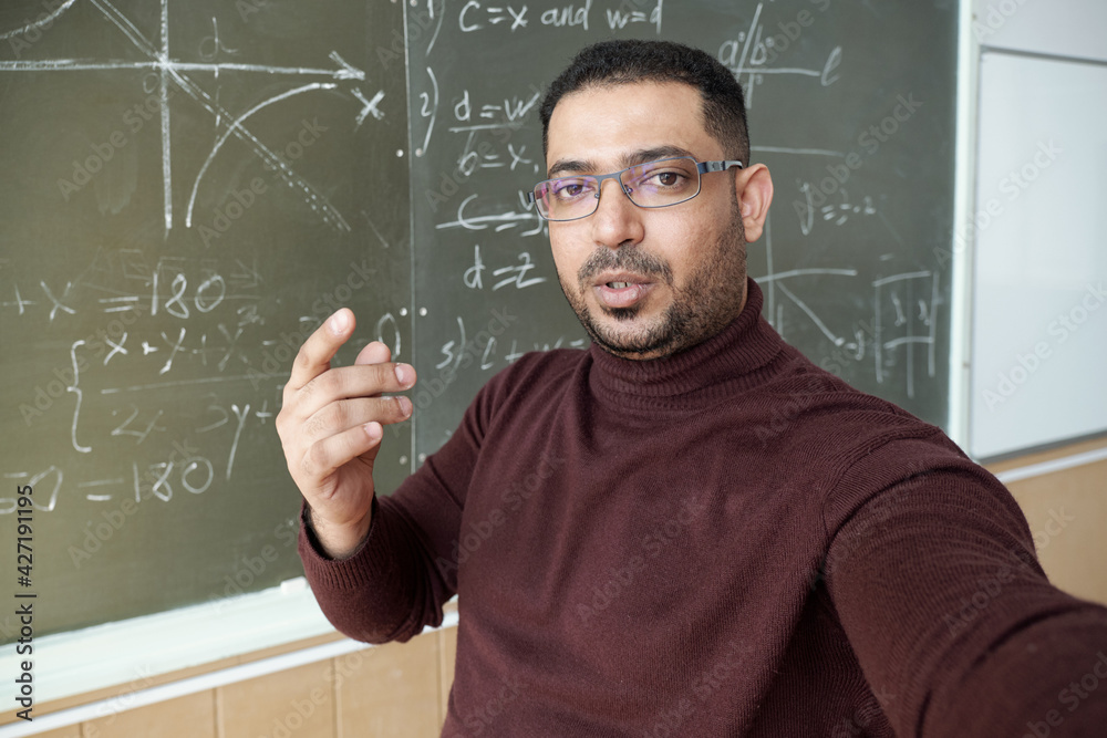 Poster Mixed-race man holding smartphone in front of himself and looking at you while standing by blackboard and explaining how to solve equation