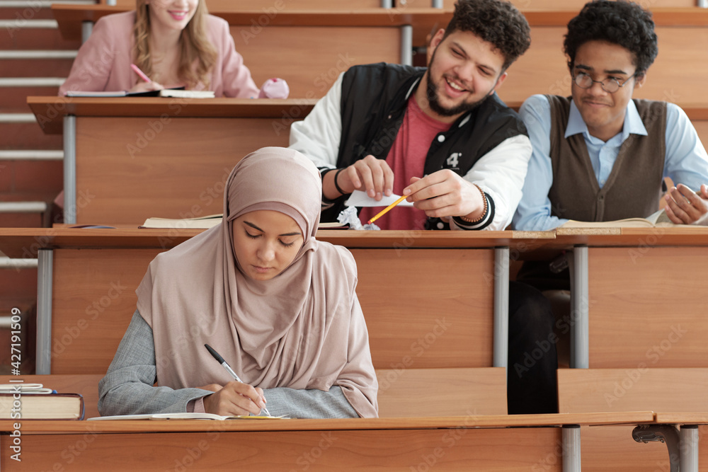 Poster serious girl in hijab making notes in copybook at lecture while sitting by desk against two intercul